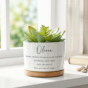 May include: A white ceramic cylindrical planter with a light wood base containing a green succulent. The planter features the name Olivia in script and the text: Some people bring so much warmth, kindness, and light into the world. You are one of them.