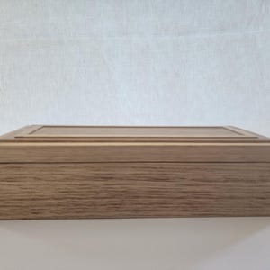 May include: A rectangular wooden box in a light brown hue. The lid features a recessed panel, and the corners are joined with dovetail joints. The wood grain is prominent, giving the box a natural appearance. The box is likely for storage.