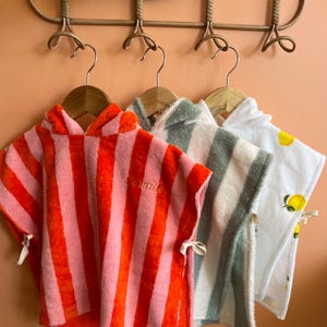 Peut inclure: Trois ponchos de plage à capuche suspendus à un porte-manteau décoratif. Les ponchos présentent différents motifs : rayures rouges et roses, rayures vertes et blanches, et blanc avec des accents de citron jaune. Chaque poncho a une capuche et des liens.