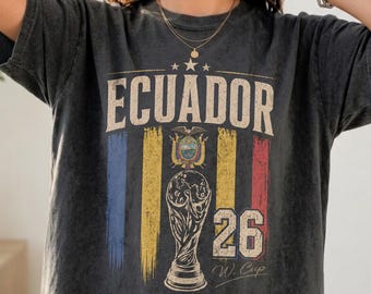 May include: A charcoal grey t-shirt featuring a distressed graphic with the text ECUADOR, a soccer trophy illustration, the number 26, and the text W. Cup Soccer Club. The design includes vertical yellow, blue, and red stripes and a small flag.
