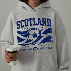 以下が含まれることがあります： 上部にSCOTLAND、中央にサッカーボール、下部にSCOTLAND SOCCER WORLD 2026の文字と小さなスコットランド国旗のアイコンが描かれた、青いグラフィックプリント付きのライトグレーのフード付きスウェットシャツ。