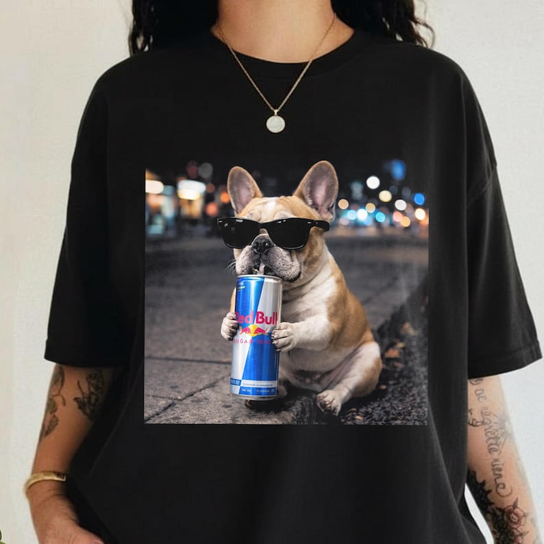 May include: Black t-shirt featuring a French bulldog wearing sunglasses and holding a Red Bull can. The dog is brown and white, set against a blurred city night scene. A gold necklace is visible.