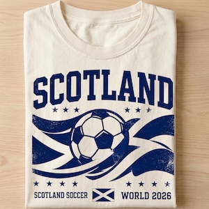 以下が含まれることがあります： クリーム色のTシャツで、太字のSCOTLANDという文字、サッカーボールのイラスト、波線、6つの星、そして下部に小さなスコットランド国旗のアイコンとSCOTLAND SOCCER WORLD 2026という文字が描かれた濃い青色のグラフィックがプリントされています。