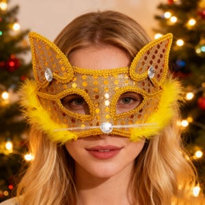 Puede incluir: Una máscara de gato dorada con lentejuelas, detalles de plumas amarillas y gemas transparentes. La máscara tiene orejas de gato y una nariz, perfecta para un disfraz o evento de máscaras. La máscara es usada por una persona con cabello rubio.