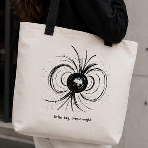 May include: A cream-coloured canvas tote bag with black fabric straps. The front features a black ink-style graphic of a central sphere with radiating curved lines and the printed text "little bag, cosmic weight" in a simple lowercase font.