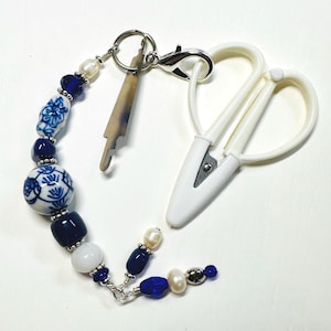May include: A white handled pair of scissors with a beaded charm. The charm features blue and white porcelain beads, dark blue beads, white beads, and pearls. A small, light brown, key-shaped charm is also attached.