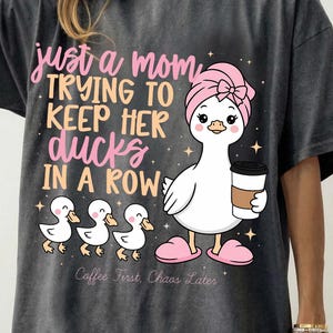 Op de afbeelding: Donkergrijze t-shirt met een cartoon eend met een roze strik en een koffiekopje, met drie eendjes. De tekst luidt "just a mom trying to keep her ducks in a row". Daaronder staat "Coffee First, Chaos Later".