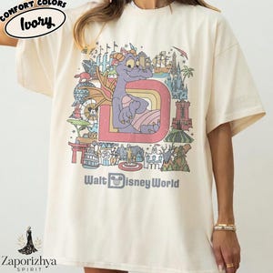 May include: Ivory-coloured t-shirt with a colourful graphic of a purple dragon inside a letter "D" with a rainbow. The graphic includes the text "Walt Disney World". The shirt has short sleeves and a relaxed fit.