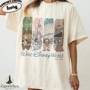 May include: Cream-coloured t-shirt with a graphic print of Walt Disney World characters and landmarks. The design includes Mickey Mouse, Minnie Mouse, Donald Duck, and Daisy Duck, with the text "Walt Disney World Est. 1971". The shirt has a loose fit.
