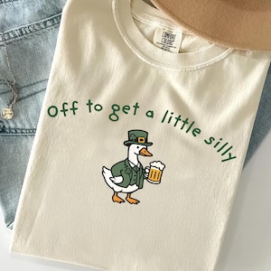 Puede incluir: Camiseta color crema con la frase "off to get a little silly" en verde. Un pato de dibujos animados con chaqueta y sombrero verdes, sosteniendo una jarra de cerveza, está impreso en la parte delantera. La camiseta está doblada sobre unos vaqueros azules.