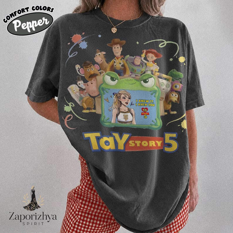 May include: A charcoal gray short-sleeve t-shirt featuring a graphic print of Toy Story characters surrounding a green frog-shaped frame. Inside the frame is an illustration of a blonde person with the text I KNEW IT, I KNEW YOU TS STORY 5.