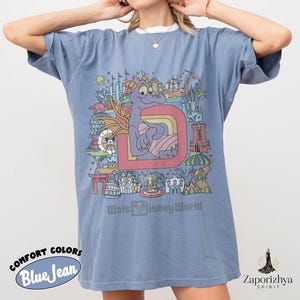 May include: Blue jean Comfort Colors t-shirt with a graphic of a purple dragon, the letter "D", and Walt Disney World landmarks. The text "Walt Disney World" is printed below. The shirt has short sleeves and a relaxed fit. The shirt is a blue colour.