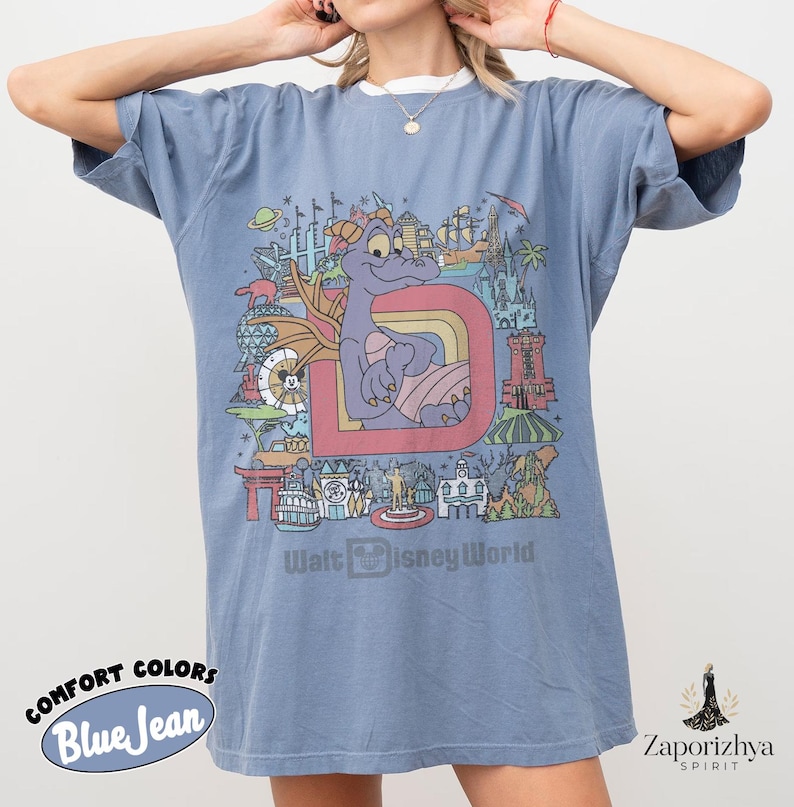May include: Blue jean Comfort Colors t-shirt with a graphic of a purple dragon, the letter "D", and Walt Disney World landmarks. The text "Walt Disney World" is printed below. The shirt has short sleeves and a relaxed fit. The shirt is a blue colour.