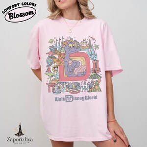 May include: Pale pink t-shirt with a colourful graphic of a purple dragon, rainbow, and landmarks. The text "Walt Disney World" is below the graphic. The shirt has the text "Comfort Colors Blossom" at the top.