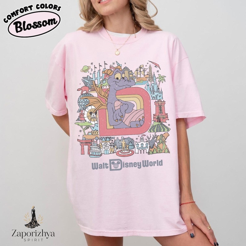 May include: Pale pink t-shirt with a colourful graphic of a purple dragon, rainbow, and landmarks. The text "Walt Disney World" is below the graphic. The shirt has the text "Comfort Colors Blossom" at the top.