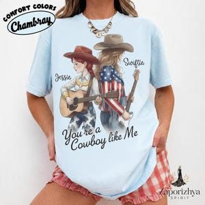 May include: A light blue t-shirt featuring a graphic of two figures in cowboy hats, one with red hair playing an acoustic guitar and one with blonde hair playing an electric guitar. The text reads Jessie, Swiftie, and You're a Cowboy like Me.