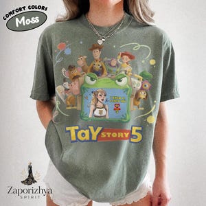 May include: A moss green short-sleeve t-shirt featuring a graphic print of various animated characters surrounding a green frame. Inside the frame is a blonde character with the text I KNEW IT I KNEW YOU and TS STORY 5. Below the frame is TAY STORY 5.