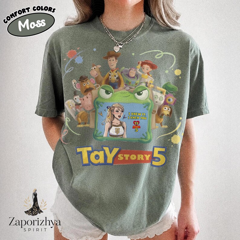 May include: A moss green short-sleeve t-shirt featuring a graphic print of various animated characters surrounding a green frame. Inside the frame is a blonde character with the text I KNEW IT I KNEW YOU and TS STORY 5. Below the frame is TAY STORY 5.