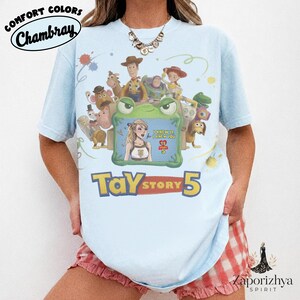 May include: A light blue t-shirt featuring a graphic print of Toy Story characters surrounding a green frog frame. Inside the frame is an illustration of a blonde person with the text I KNEW IT I KNEW YOU TS STORY 5. Below the graphic is the text TAY STORY 5.
