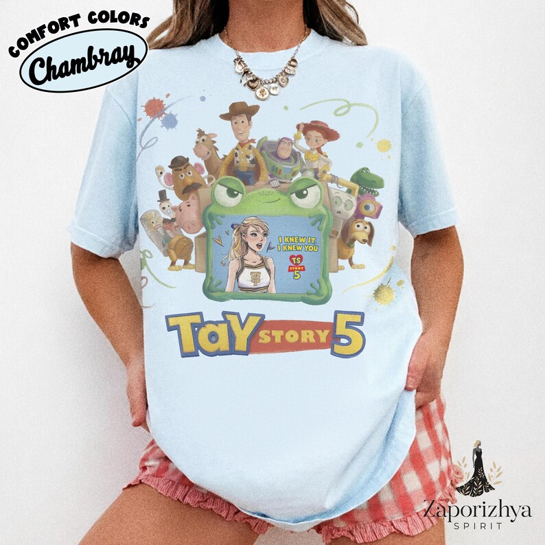 May include: A light blue t-shirt featuring a graphic print of Toy Story characters surrounding a green frog frame. Inside the frame is an illustration of a blonde person with the text I KNEW IT I KNEW YOU TS STORY 5. Below the graphic is the text TAY STORY 5.