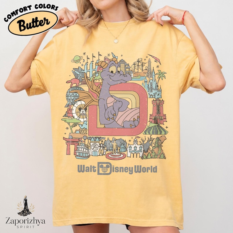 May include: Yellow t-shirt with a colourful graphic featuring a purple dragon inside a large letter "D" and various Disney World landmarks. The text "Walt Disney World" is printed below the graphic. The shirt has the text "Comfort Colors Butter" in a circle at the top.