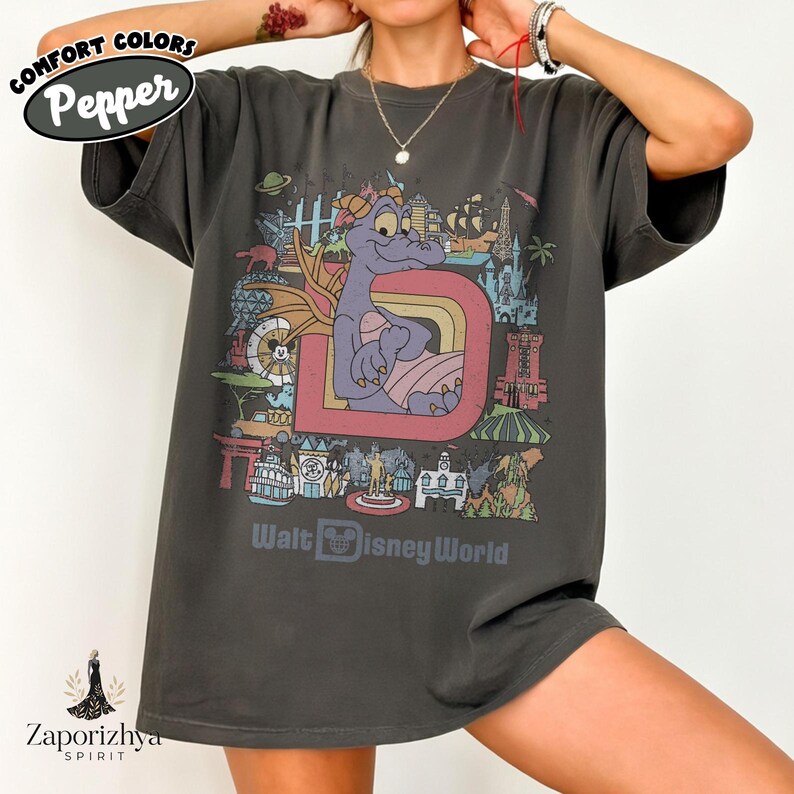 May include: Dark grey t-shirt with a colourful graphic of a purple dragon character inside a large letter "D" with a rainbow. The shirt also includes the text "Walt Disney World" and the Comfort Colors and Zaporizhya Spirit logos.