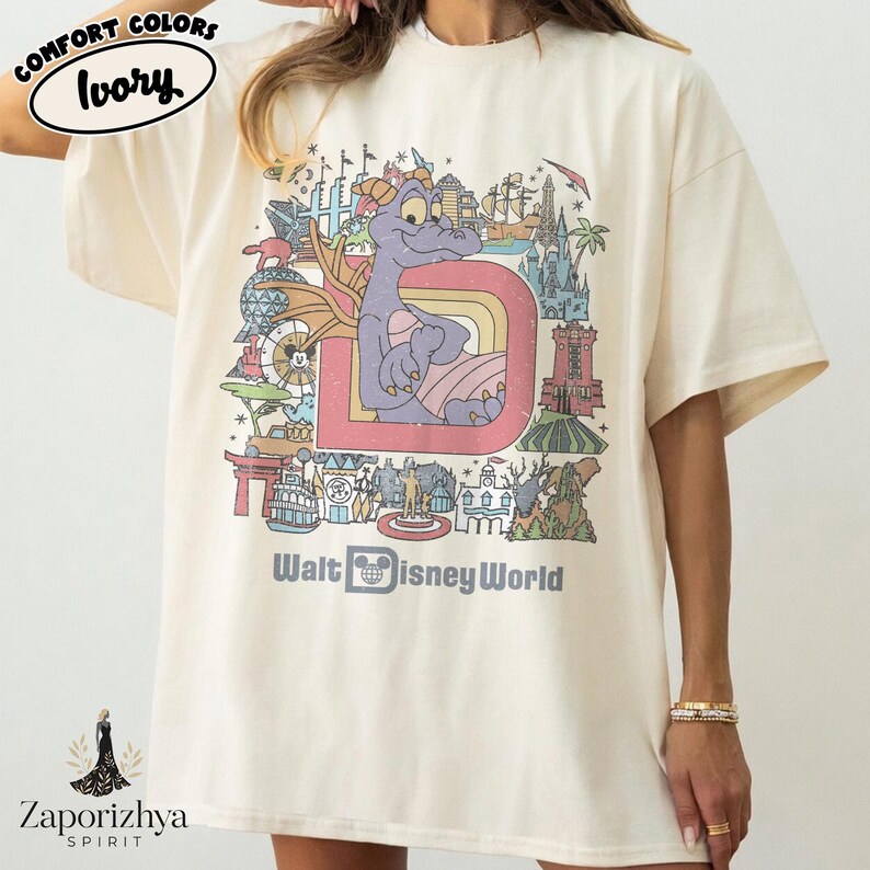 May include: Ivory-coloured t-shirt with a colourful graphic of a purple dragon inside a letter "D" with a rainbow. The graphic includes the text "Walt Disney World". The shirt has short sleeves and a relaxed fit.