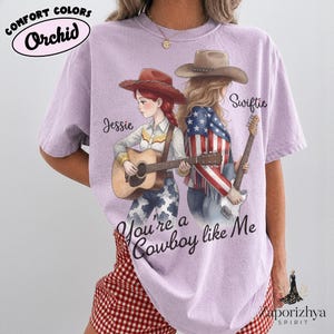 May include: A light purple t-shirt featuring a graphic of two figures in cowboy hats. One figure plays an acoustic guitar, the other an electric guitar. Text reads Jessie, Swiftie, and You're a Cowboy like Me. The shirt is labeled Comfort Colors Orchid.