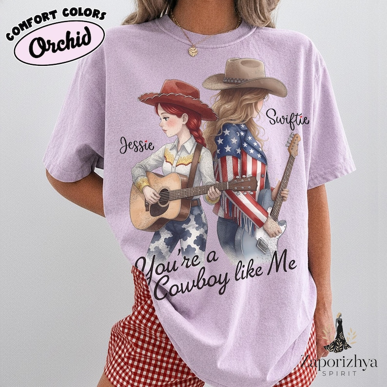 May include: A light purple t-shirt featuring a graphic of two figures in cowboy hats. One figure plays an acoustic guitar, the other an electric guitar. Text reads Jessie, Swiftie, and You're a Cowboy like Me. The shirt is labeled Comfort Colors Orchid.