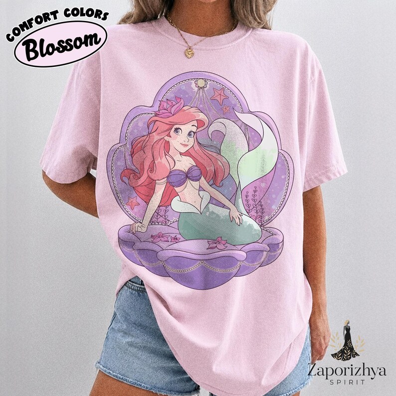 May include: A light pink t-shirt featuring a graphic of a mermaid with long red hair and a green tail, sitting inside a large purple seashell. The design includes small stars and floral accents. The text COMFORT COLORS Blossom is in the top left corner.