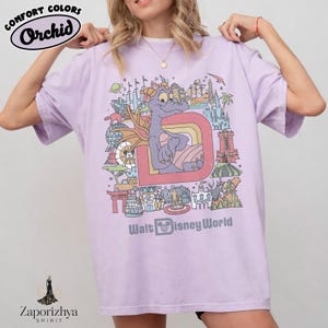 May include: A lilac t-shirt with a colourful graphic featuring a purple dragon character, the letter "D", and various landmarks. The text "Walt Disney World" is printed below the graphic. The shirt also has the text "Comfort Colors" and "Orchid".
