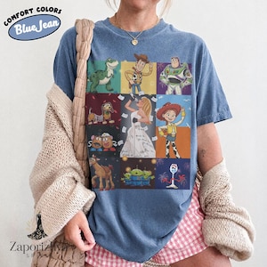 May include: A blue jean colored t-shirt featuring a grid of nine panels displaying various animated characters including a dinosaur, a cowboy, a space ranger, a dog, a singing figure, a cowgirl, potato figures, aliens, and a white spork character.
