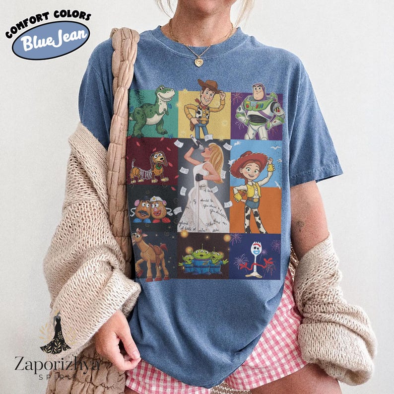 May include: A blue jean colored t-shirt featuring a grid of nine panels displaying various animated characters including a dinosaur, a cowboy, a space ranger, a dog, a singing figure, a cowgirl, potato figures, aliens, and a white spork character.