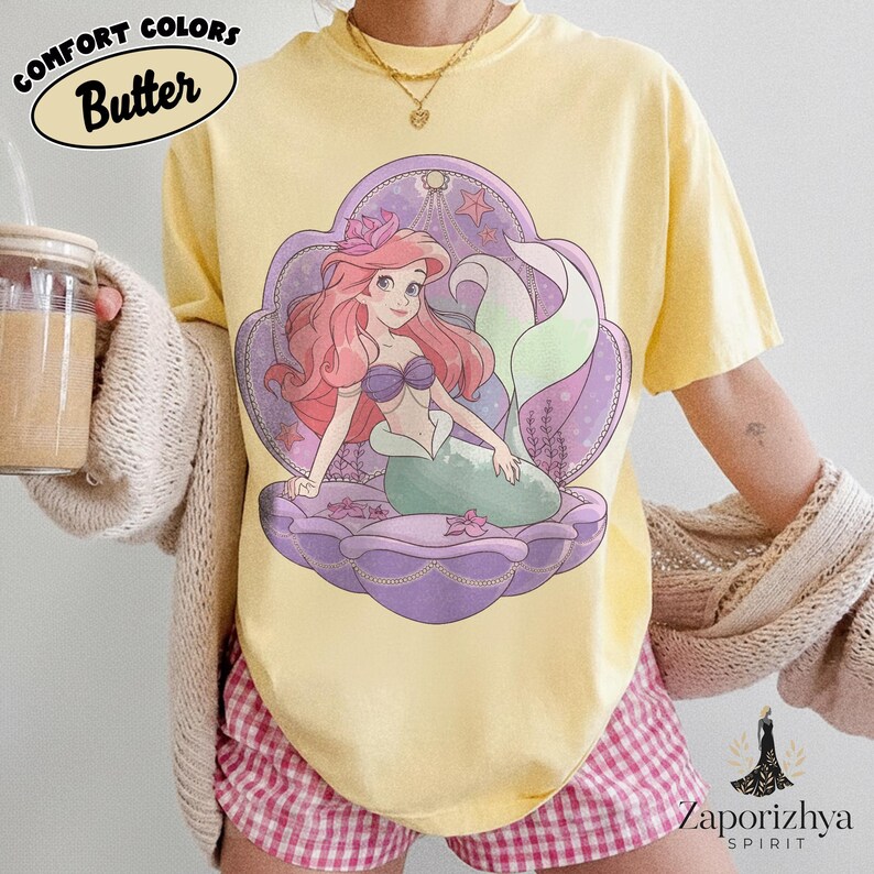 May include: A butter yellow short-sleeve t-shirt featuring a graphic of a mermaid with long pink hair and a green tail, sitting inside a large purple seashell. The design includes small pink flowers and stars, with the text Comfort Colors Butter.