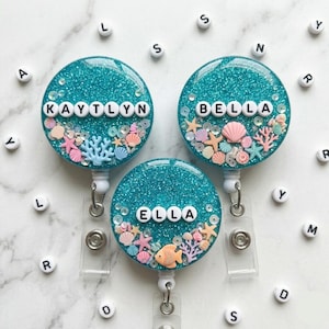 May include: Three teal glitter badge reels with the names "Kaytlyn", "Bella", and "Ella" spelled out in white letters. Each reel is decorated with miniature seashells, starfish, and coral. The reels have a clear plastic strap and a metal clip.