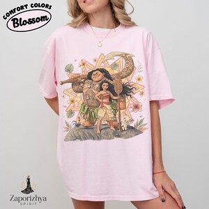 May include: Light pink t-shirt featuring a colorful graphic of characters from a popular animated movie. The design includes a large figure with tattoos, a young woman, and a small pig. The shirt has the words "Comfort Colors Blossom" in a black oval.