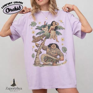 Może przedstawiać: Jasnofioletowa koszulka z grafiką postaci z kresk&oacute;wek. Projekt zawiera palmę, kwiaty i kilka postaci. Koszulka ma u g&oacute;ry napis "Comfort Colors Orchid". Marka "Zaporizhya Spirit" znajduje się na dole.