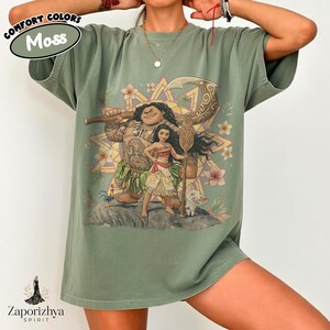 May include: A moss green t-shirt featuring a colorful graphic of characters from the animated film "Moana." The design includes Maui, Moana, and a small pig. The shirt has the text "Comfort Colors Moss" and the brand logo "Zaporizhya Spirit."