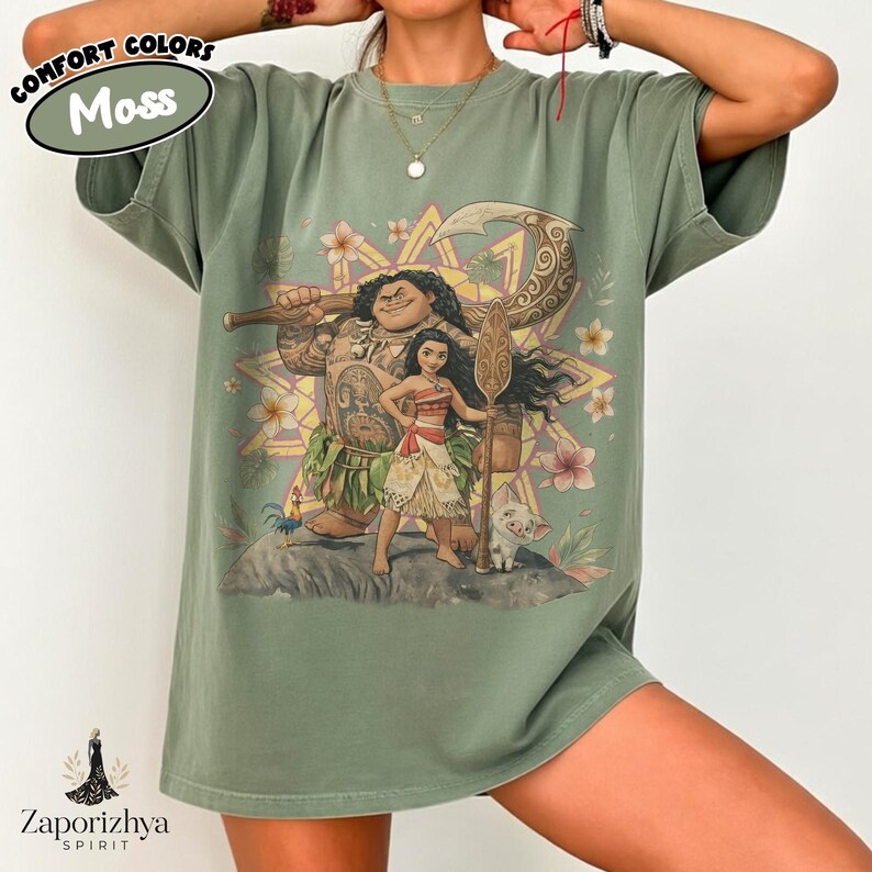 May include: A moss green t-shirt featuring a colorful graphic of characters from the animated film "Moana." The design includes Maui, Moana, and a small pig. The shirt has the text "Comfort Colors Moss" and the brand logo "Zaporizhya Spirit."