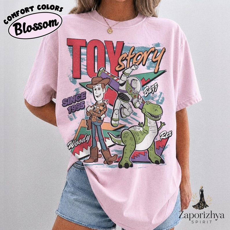 May include: A light pink short-sleeve t-shirt featuring a graphic print of Toy Story characters Woody, Buzz Lightyear, and Rex. The design includes the text "TOY story", "SINCE 1995", "Woody", "Buzz", and "Rex" with colorful geometric shapes.