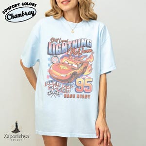 May include: Light blue t-shirt featuring a graphic of Lightning McQueen from the movie Cars. The design includes the text "Street Legend Lightning McQueen", "Need for Speed", and the number "95". The shirt is from Comfort Colors.