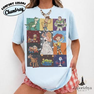 May include: A light blue t-shirt featuring a grid of nine panels displaying various animated characters, including a dinosaur, a cowboy, a space ranger, a dog, a singer in a white dress, a cowgirl, potato figures, a horse, aliens, and a fork character.