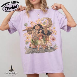 May include: A light purple t-shirt featuring a graphic of characters from a popular animated movie. The design includes a large figure holding a weapon, a young woman, and a small pig. The shirt has the text "Comfort Colors Orchid" at the top.