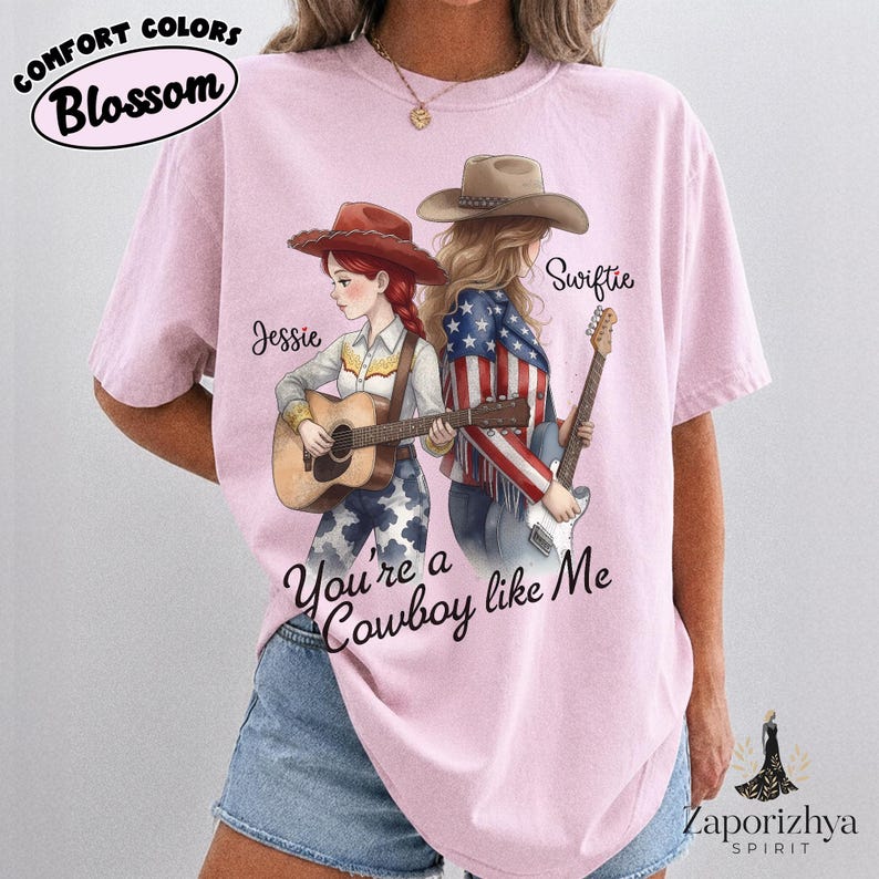 May include: A light pink short-sleeve t-shirt featuring a graphic of two figures in cowboy hats, one with red hair playing an acoustic guitar and one with blonde hair playing an electric guitar. The text reads Jessie, Swiftie, and You're a Cowboy like Me.