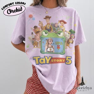 May include: A light purple short-sleeve t-shirt featuring a graphic print of Toy Story characters surrounding a green frog-shaped frame. Inside the frame is an illustration of a blonde person with the text I KNEW IT I KNEW YOU TS STORY 5.