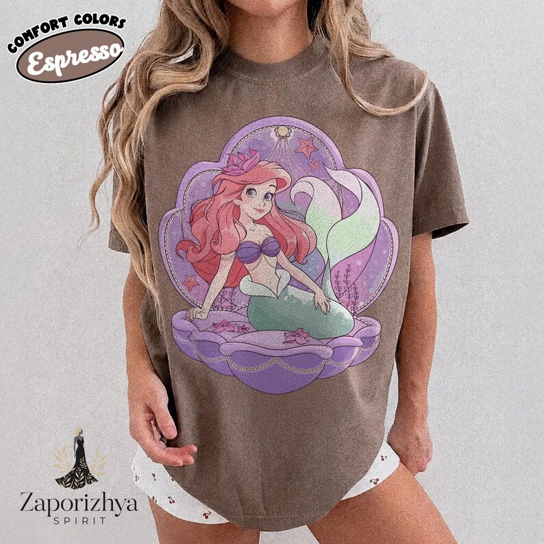 May include: An espresso brown short-sleeve t-shirt featuring a graphic of a mermaid with long red hair and a green tail sitting inside a large purple seashell. The design includes small pink flowers and stars. Text on the shirt reads COMFORT COLORS Espresso.