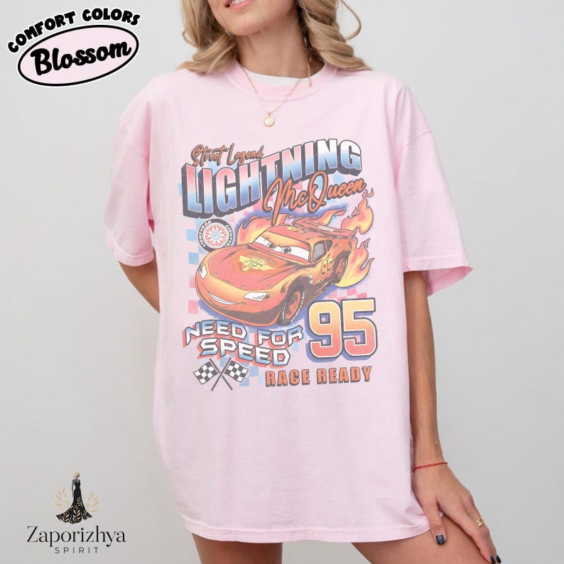 May include: Light pink t-shirt featuring a graphic of Lightning McQueen from the movie Cars. The design includes the text "Street Legends Lightning McQueen 95 Need for Speed Race Ready". The shirt has short sleeves and a relaxed fit.