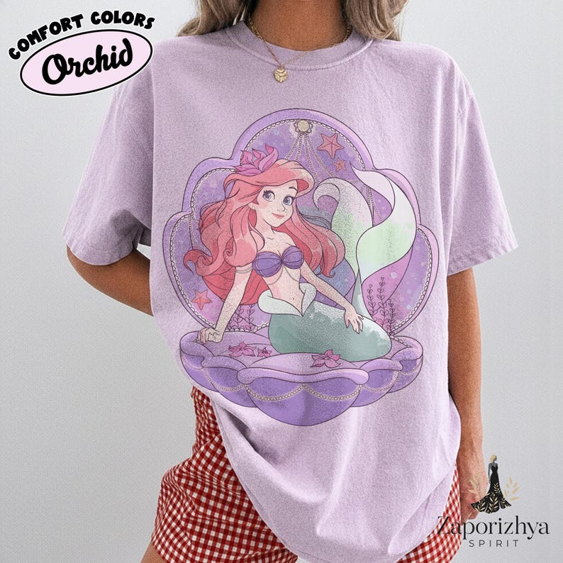 May include: A light purple short-sleeve t-shirt featuring a graphic of a mermaid with long red hair and a green tail sitting inside a large purple seashell. The text COMFORT COLORS Orchid is printed in the upper left corner of the garment.