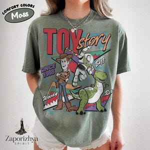 May include: A moss green short-sleeve t-shirt featuring a graphic print of Toy Story characters Woody, Buzz Lightyear, and Rex. The design includes the text TOY story, SINCE 1995, Woody, Buzz, and Rex, set against a geometric background pattern.