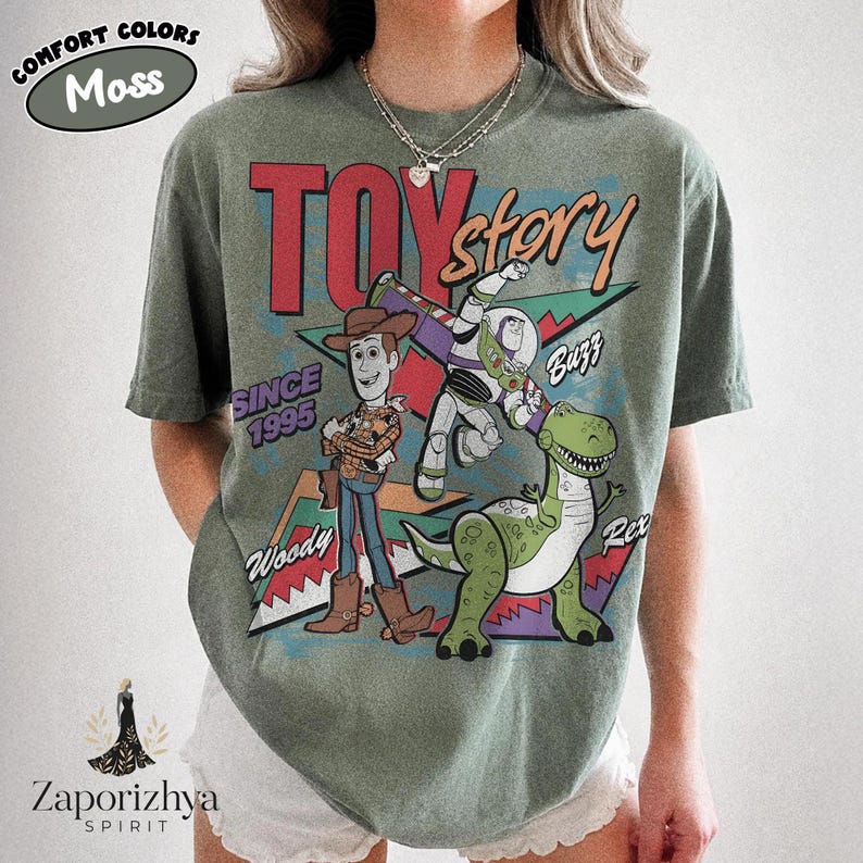 May include: A moss green short-sleeve t-shirt featuring a graphic print of Toy Story characters Woody, Buzz Lightyear, and Rex. The design includes the text TOY story, SINCE 1995, Woody, Buzz, and Rex, set against a geometric background pattern.